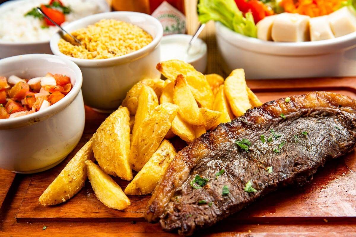 Fotografia de carne grelhada com batatas fritas, farofa e vinagrete